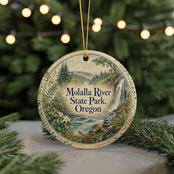 A round ornament with a scenic landscape of the Molalla River State Park in Oregon, featuring a waterfall and pine trees, is displayed on a table with a blurred background of Christmas lights.