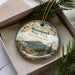 A decorative ornament in a box, featuring a scenic mountain landscape with pine trees and a sign that says "Monadnock State Park, New Hampshire".