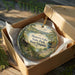 An ornament in a box, featuring a scenic landscape with mountains and a cabin, with the text "Moncover Lake State Park, West Virginia" printed on it.