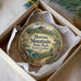 A decorative ornament in a white box, featuring a scenic landscape with mountains, trees, and a bird.