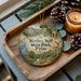 A decorative ornament with a picture of a park, featuring a bird, flowers, and a path, sits on a wooden tray next to a lit candle and pine cones.