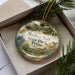 A decorative ornament in a box, featuring a scenic landscape with a house, trees, and wildlife.