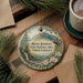 A decorative ornament featuring a mountain landscape with a waterfall, surrounded by pine branches and flowers, placed on a wooden surface next to a cup of coffee.