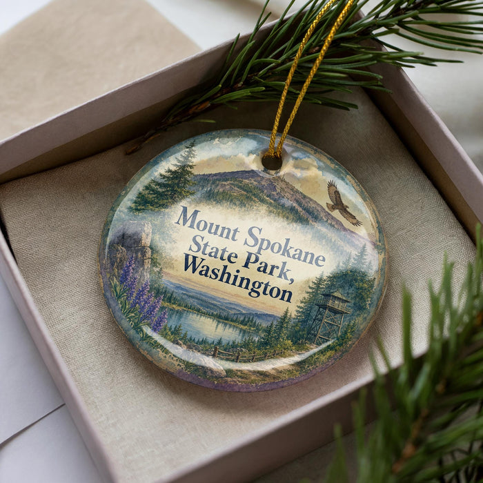 A decorative ornament in a box, featuring a scenic landscape with mountains, pine trees, and a lake.