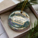 A decorative ornament in a box, featuring a scenic landscape with mountains, pine trees, and a lake.