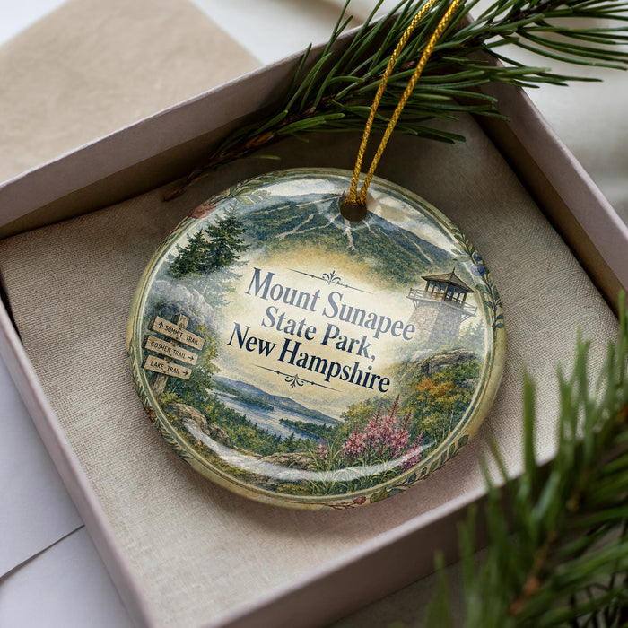 A decorative ornament in a box, featuring a scenic landscape with mountains, pine trees, and a lighthouse, with the text "Mount Sunapee State Park, New Hampshire" prominently displayed.