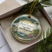 A decorative ornament in a box, featuring a scenic landscape with mountains, pine trees, and a lighthouse, with the text "Mount Sunapee State Park, New Hampshire" prominently displayed.