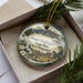 A decorative ornament featuring a scenic landscape of Mount Washington State Park, New Hampshire, with a mountain, pine trees, and a signpost.