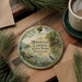 A decorative ornament featuring a scenic image of a lake and a cabin, with the text "Mousetail Landing State Park, Tennessee" printed on it. The ornament is placed on a wooden surface, surrounded by pine branches and a cup of coffee.