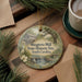 A decorative ornament featuring a scenic image of Musgrove Mill, a historic site in South Carolina, surrounded by pine branches and a cup of coffee.