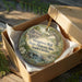 A wooden box containing a decorative ornament with a scenic image of Musgrove Mill, South Carolina.