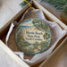 A circular ornament with a beach scene and text, placed in a box with a white tissue paper lining.
