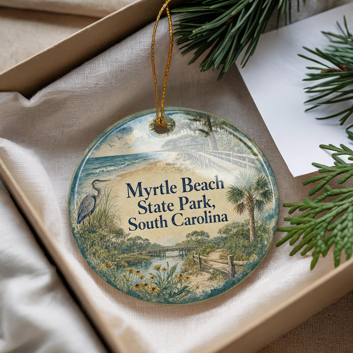 A decorative ornament with a beach scene and text, placed in a box with pine branches.