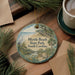 A decorative ornament featuring a scenic beach landscape with a bird, palm trees, and a wooden pier, placed on a wooden surface alongside a cup of coffee and some pine branches.