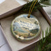 A decorative ornament in a box, featuring a scenic landscape with mountains, trees, and a body of water.
