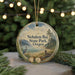 A round ornament with a scenic landscape of a lake, mountains, and trees, featuring a bird and a heron, is displayed on a table with a blurred background of a Christmas tree and lights.