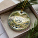 A decorative ornament in a box, featuring a scenic landscape with a bird and waterfalls, surrounded by pine branches.