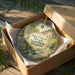 A decorative ornament in a brown box, featuring a scenic landscape with a waterfall and greenery, with the text "New Glarus Woods State Park Wisconsin" printed on it.