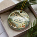 A decorative ornament in a box, featuring a scenic landscape with a river and mountains, along with the text "Newport State Park, Wisconsin".