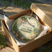 A decorative ornament in a brown box, featuring a scenic landscape with a river and trees, with the text "Newport State Park, Wisconsin" printed on it.