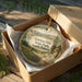 A round ornament in a brown box, featuring a scenic landscape with mountains, trees, and a winding path, with the text "Newton Hills State Park, South Dakota" printed on it.