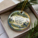 A decorative ornament in a box, featuring a scenic landscape with a cannon, trees, and a state park.