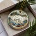 A decorative ornament with a scenic lake and forest landscape, featuring a cabin and a bird, is displayed in a box with pine branches.