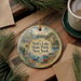 A round ornament with a scenic lake and forest landscape, featuring a duck and boat, is displayed on a wooden surface alongside a cup of coffee and some pine branches.