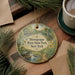 A decorative ornament with a scenic illustration of a river and park, featuring a bird and a turtle, is displayed on a wooden surface alongside a cup of coffee and some papers.