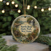 A decorative ornament featuring a scenic landscape with a waterfall, a cabin, and a bridge, hanging in front of a Christmas tree with lights.