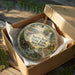 A decorative ornament in a box, featuring a scenic landscape with a waterfall and a small cabin, with the text "North Bend State Park, West Virginia" printed on it.