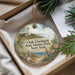 A decorative ornament with a lighthouse and a coastal scene, placed in a box with pine branches.