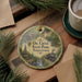 A decorative ornament featuring illustrations of Pennsylvania State Park, including a train, a waterfall, and a cabin, is displayed on a wooden surface alongside a cup of coffee and some pine branches.