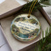 A decorative ornament with a scenic image of Okmulgee Springs State Park, Oklahoma, displayed in a box with pine branches.