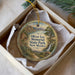 A round ornament with a landscape scene and text, placed in a box with a white tissue paper lining.