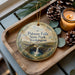 A decorative ornament with a scenic image of Palouse Falls State Park, Washington, placed on a wooden tray alongside pine cones and a lit candle.