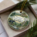 A decorative ornament in a box, featuring a scenic landscape with a black bear and a waterfall.
