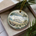 A decorative ornament in a box, featuring a scenic landscape with mountains, pine trees, and a body of water.