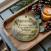 A decorative ornament with a landscape scene and text, placed on a wooden surface next to pine cones and a lit candle.