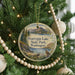 A round ornament hanging on a Christmas tree, featuring a scenic landscape with mountains, trees, and a bird.
