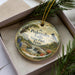 A decorative ornament in a box, featuring a scenic landscape with a waterfall and pine trees.