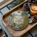A decorative ornament with a scenic forest landscape, featuring a waterfall, pine trees, and a deer, is displayed on a wooden tray alongside pine cones and a lit candle.