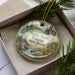 A decorative ornament in a box, featuring a scenic landscape with a forest, a waterfall, and a deer.