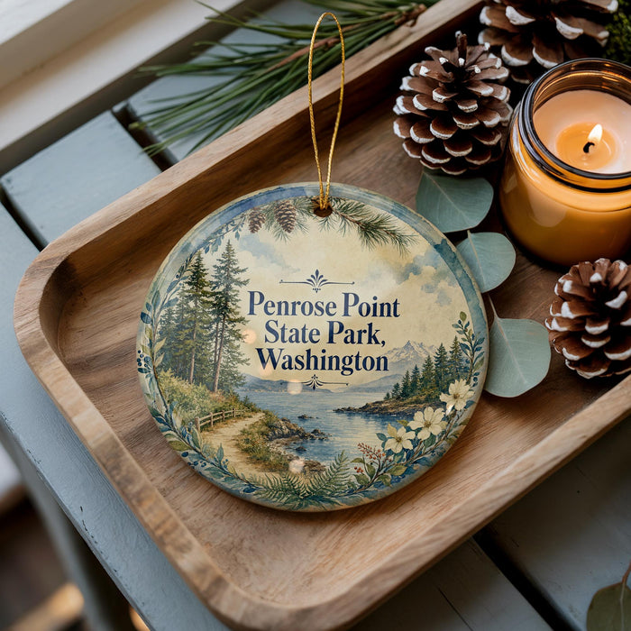 A decorative ornament with a scenic landscape of Penrose Point State Park, Washington, featuring pine trees, a lake, and mountains. The ornament is displayed on a wooden tray, accompanied by a lit candle and pine cones.
