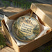 A decorative ornament in a wooden box, featuring a scenic landscape with mountains and pine trees.