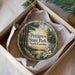 A decorative ornament in a box, featuring a scenic landscape with a wooden bridge, trees, and a sunset.