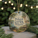 A round ornament with a painting of a scenic landscape, featuring a river, trees, and a cabin, hanging in front of a blurred Christmas tree with lights.
