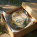 A circular ornament in a wooden box, featuring a scenic landscape with mountains and a river, with the text "Pictograph Cave State Park, Montana" printed on it.