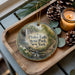 A wooden tray with a decorative ornament featuring a scenic image of Pixley Falls State Park in New York, accompanied by pine cones and a lit candle.