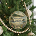 A round ornament hanging on a Christmas tree, featuring a scenic landscape with a lighthouse and a body of water.